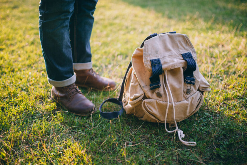 A person wearing rolled-up jeans and brown boots stands on grass next to a tan canvas backpack with black straps and white drawstrings.