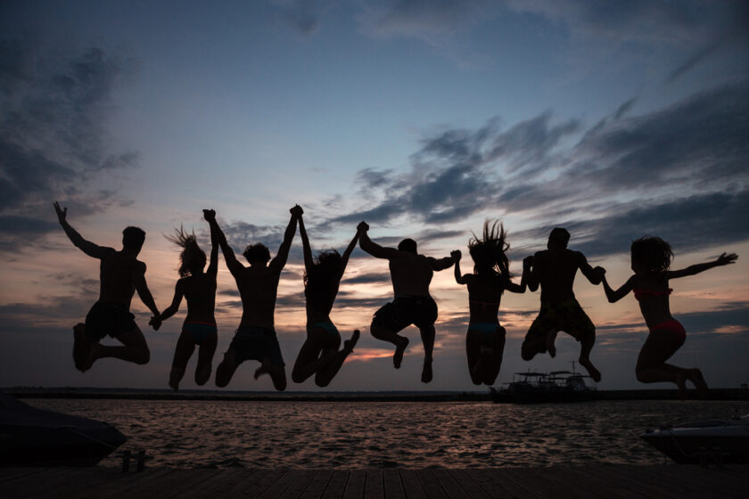 Silhouettes of people jumping with arms raised against a sunset sky over water. They are on a wooden dock, with boats visible in the background. The sky features dramatic clouds with shades of orange, pink, and blue.