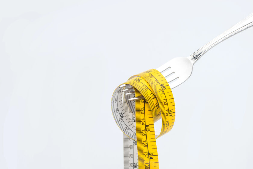 A fork holds a coiled yellow measuring tape against a plain white background, symbolizing dieting or portion control.