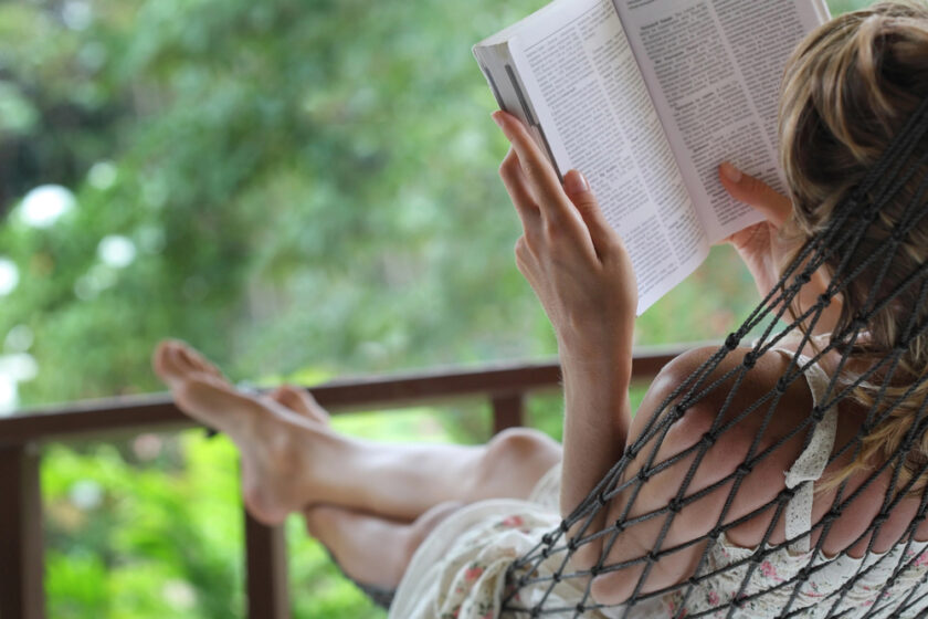 A person relaxes in a hammock on a porch, reading a book. Their legs are crossed, and they are surrounded by lush green foliage, creating a peaceful and tranquil atmosphere.