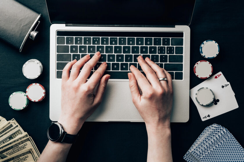 A person types on a laptop surrounded by poker chips, playing cards, a flask, and stacks of cash, suggesting online gambling or poker playing.