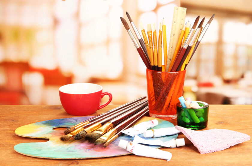 A colorful collection of art supplies on a wooden table, including paintbrushes in a red container, a paint palette, tubes of paint, a red cup, chalk pieces in a green holder, and a pink cloth, set against a blurred background.