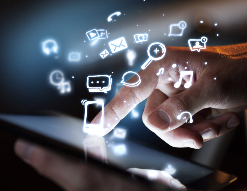 A hand touches a tablet screen, with glowing digital icons such as email, music, chat, and search floating above, representing connectivity and digital technology.