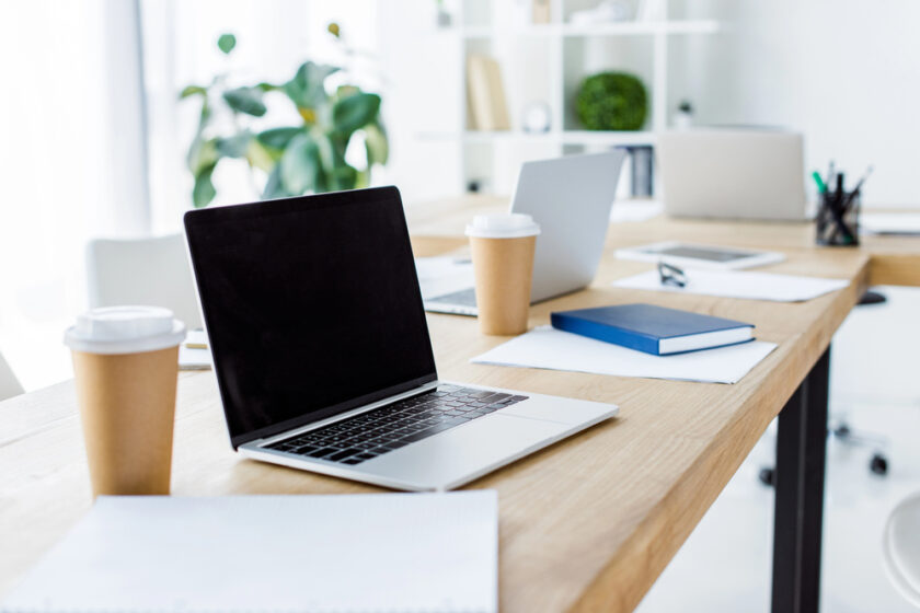 Modern office workspace with open laptops, notebooks, papers, and disposable coffee cups on a wooden desk; shelves and a green plant are in the background, suggesting a clean, organized environment.