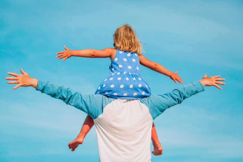 A young girl in a blue polka dot dress sits on an adult’s shoulders, spreading her arms wide. The adult wears a white shirt with blue sleeves. They face a bright blue sky, seen from behind.