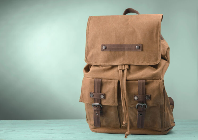 A brown canvas backpack with leather straps and multiple pockets sits on a light turquoise wooden surface against a gradient green background.