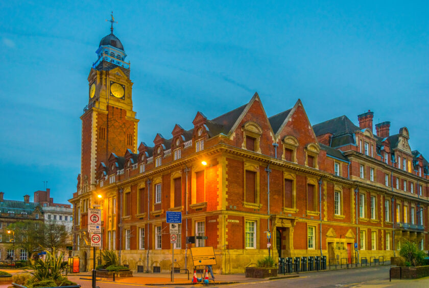 A historic red-brick building with a tall clock tower stands on a street corner at dusk, illuminated by streetlights against a blue sky.