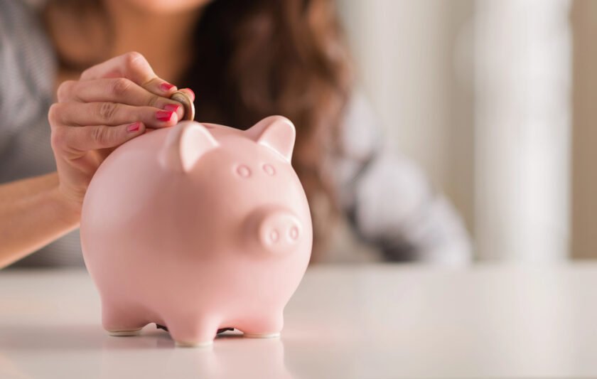 A person with painted nails is inserting a coin into a pink piggy bank. The background is blurred, and the focus is on the hand and the piggy bank placed on a white surface.