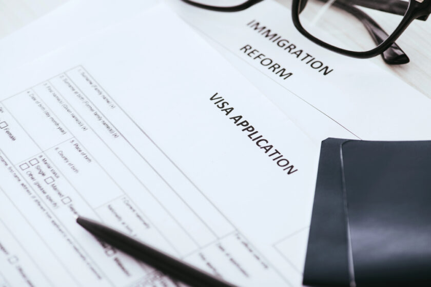 A close-up of a visa application form, a document labeled “Immigration Reform,” a pen, a pair of eyeglasses, and a dark-colored passport on a desk.