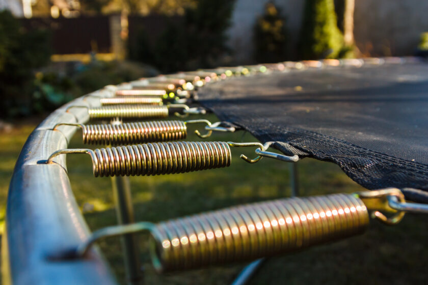 Close-up of a trampolines metal springs attached to its metal frame and black jumping mat. The springs are arranged in a row, stretching into the distance. The background is a blurred garden with trees and a fence.