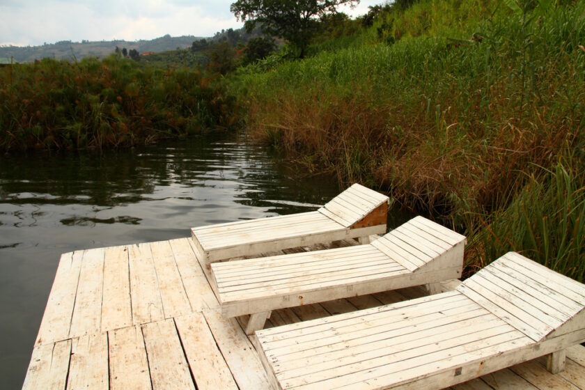 Two white wooden loungers are positioned on a wooden deck extending over a calm lake. The deck is surrounded by lush greenery and tall grasses, with hills visible in the background under a cloudy sky.