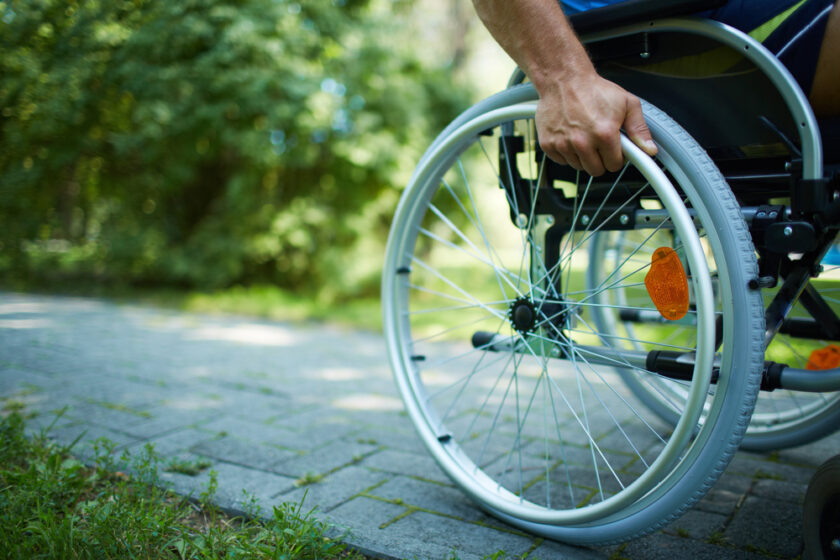 A person in a wheelchair is moving along a paved path in a park, with one hand on the wheel. The background features lush green foliage.