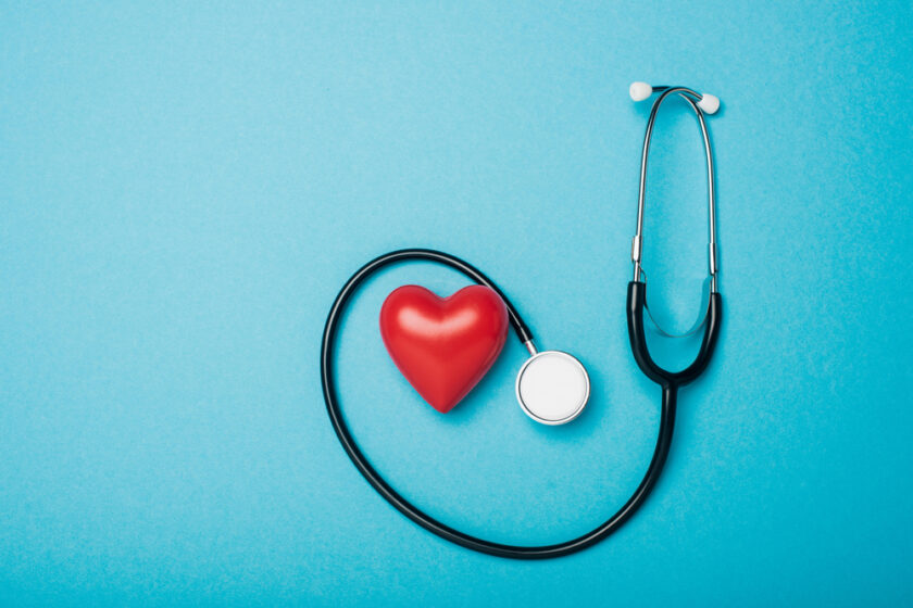 A stethoscope with its tubing shaped like a heart surrounds a red heart model on a blue background, symbolizing heart health and medical care.