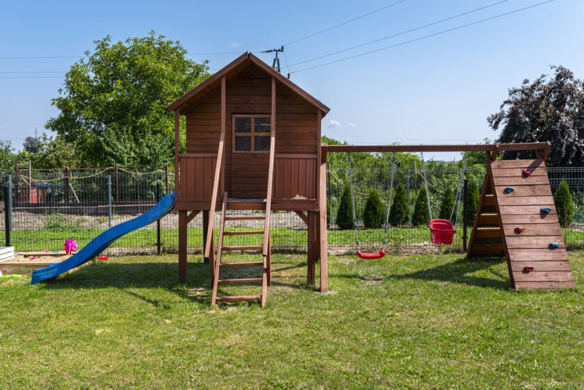 A wooden playhouse on stilts with a slide, ladder, and climbing wall stands on a grassy area. It is surrounded by a fence and trees in the background. A red toy is visible nearby. The sky is clear and blue.