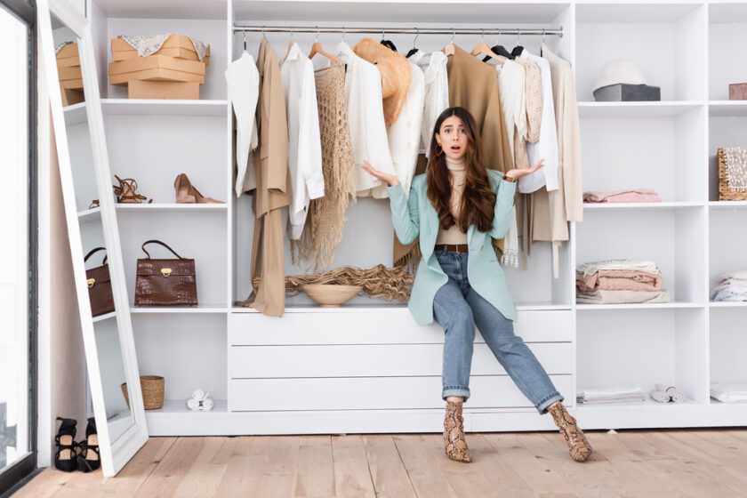 A young woman sits in front of an open wardrobe, shrugging with her palms up. She is surrounded by neatly organized, neutral-toned clothes and accessories. The room appears bright and modern.