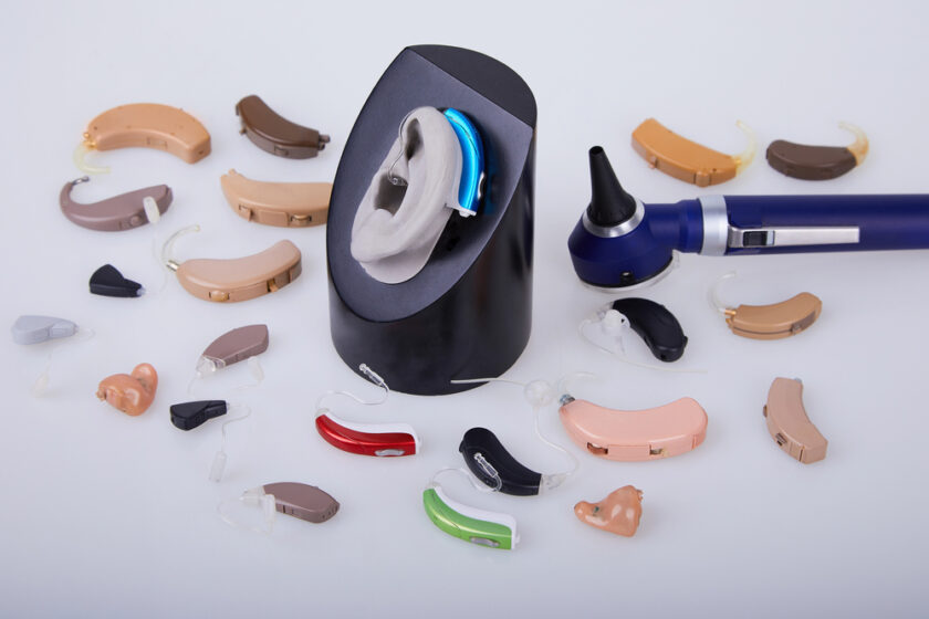 Various hearing aids surround a central display with an ear model wearing a blue hearing aid. An otoscope is placed on the right. The hearing aids come in different sizes and colors, including beige, brown, and red.