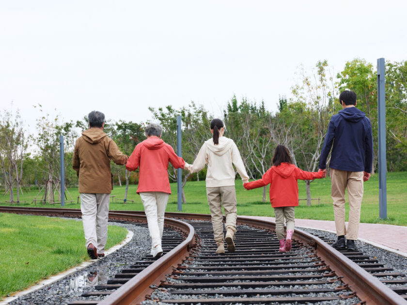 Five people, including two children and three adults, walk hand-in-hand along train tracks in a park setting. They are dressed in casual jackets and pants, with greenery and trees visible in the background. The sky is overcast.