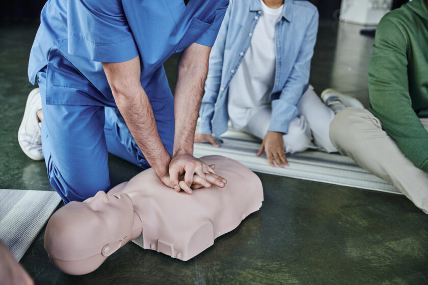 A person in blue scrubs demonstrates CPR on a mannequin, pressing on its chest. Two others are seated nearby, observing on mats.