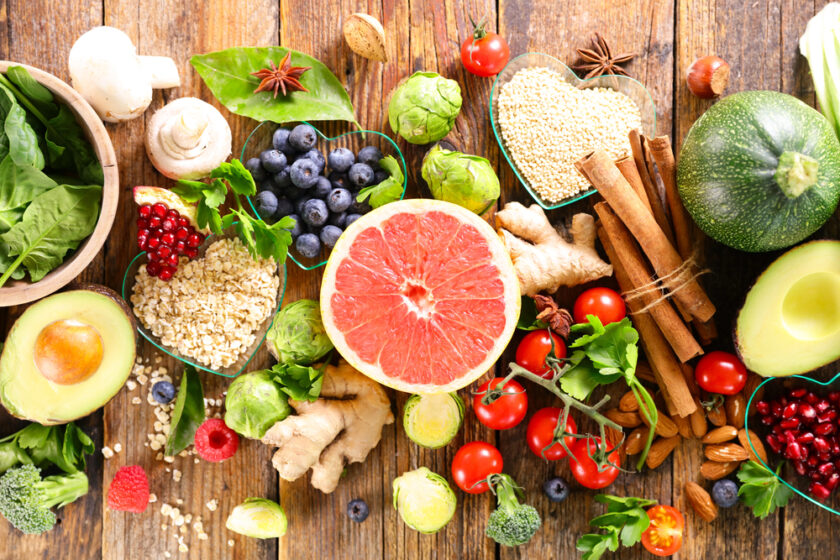 A colorful assortment of fresh fruits, vegetables, grains, and spices, including grapefruit, avocado, berries, tomatoes, spinach, ginger, Brussels sprouts, oats, and cinnamon sticks, arranged on a rustic wooden table.