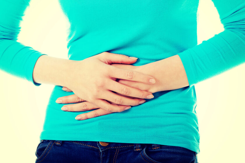 A person wearing a turquoise long-sleeve shirt and jeans holds their hands over their stomach, suggesting discomfort or abdominal pain.