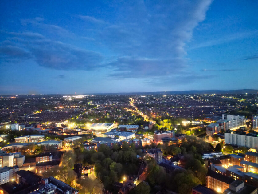 Aerial view of a city at dusk with illuminated buildings, glowing streetlights, and dark green trees, under a blue sky with scattered clouds stretching across the horizon.