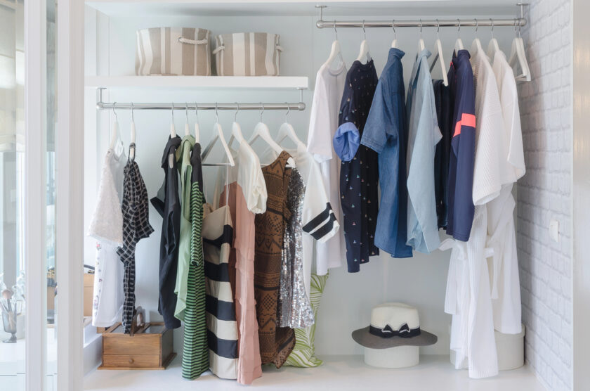 A neatly organized open closet with various shirts, blouses, and dresses hanging on two racks, a striped storage basket on a shelf, a wooden box, and a white hat with a black ribbon on the bottom shelf.