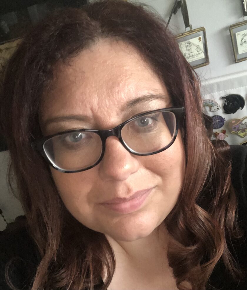 Framed pictures adorn the wall behind, adding a personal touch to the scene as a person with long brown hair and glasses takes a selfie. Their dark top contrasts with their bright smile, providing a perfect glimpse into their about me moment indoors.
