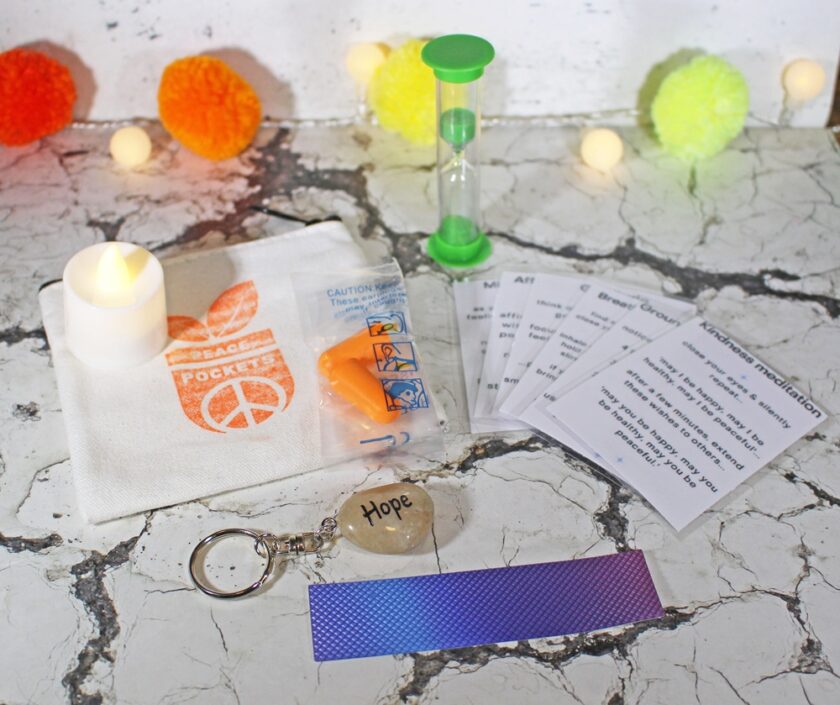 A collection of items including a stone with Hope inscribed, a peace sign pouch, cards with messages, a green hourglass, orange earplugs, a purple reflective strip, and an LED tealight, on a marble surface with colorful pom-poms in the background.