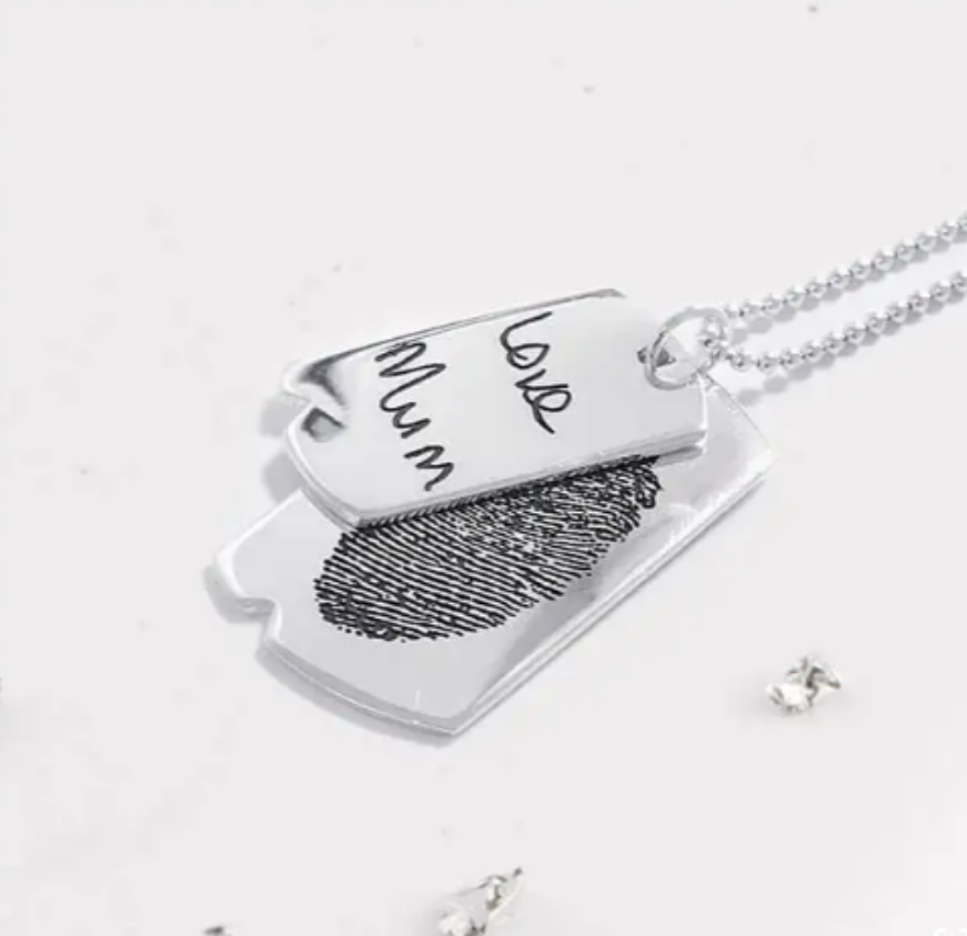 Silver layered pendant necklace featuring a fingerprint design on one tag and handwritten text saying love Mum on the other. The necklace rests on a white surface scattered with small metallic pieces.
