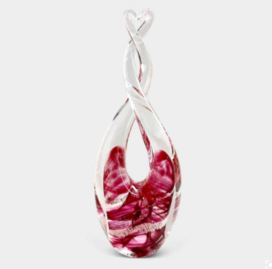 A glass sculpture with an abstract swirling design, featuring intertwined clear and red glass elements. The piece has a smooth, elongated form with a heart-like shape at the top. The background is plain white.