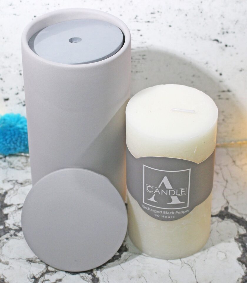 A white pillar candle labeled Candle A stands next to a gray cylindrical container with its matching lid resting in front on a marbled surface. A blue fuzzy object is partially visible in the background.