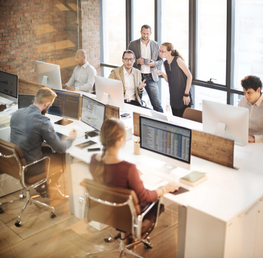 Modern open-plan office with several people working at computers. Three colleagues stand and talk near large windows, while others focus on their monitors at long shared desks. Exposed brick walls and natural light fill the space.