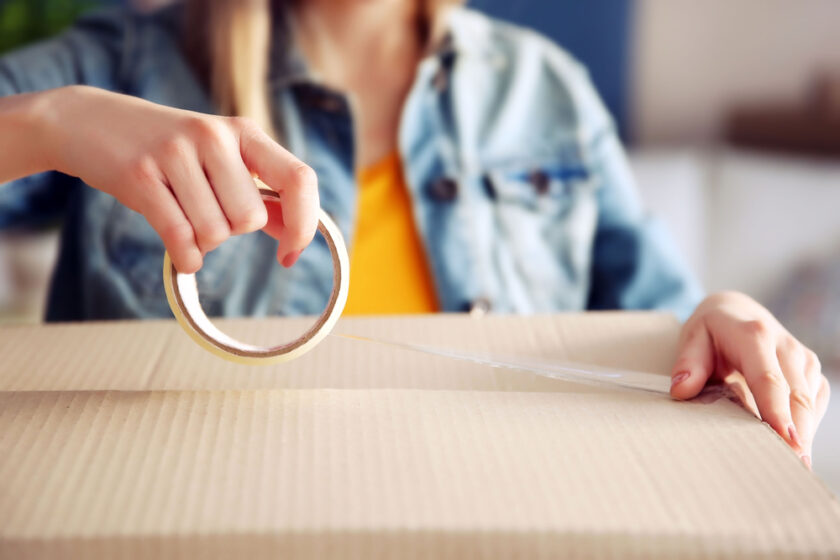 A person in a denim jacket seals a cardboard box with clear packing tape, suggesting packing or moving activities.