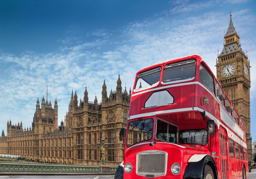 A classic red double-decker bus in front of the Houses of Parliament and Big Ben under a partly cloudy blue sky in London.