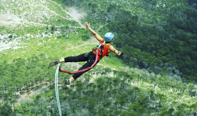 A person wearing a helmet and harness is bungee jumping off a high platform surrounded by lush green forest and rocky terrain below.