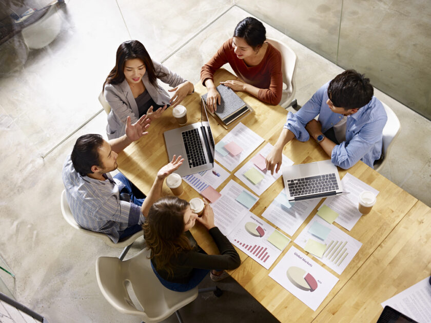 Five people sit around a wooden table with laptops, papers, and coffee cups, discussing graphs and charts. The image is taken from above in a bright, modern office setting.