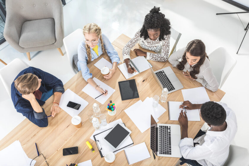 Five people sit around a wooden table with laptops, notebooks, and papers, working and discussing together. Some are writing while others look at screens. Coffee cups, pens, and office supplies are scattered on the table.