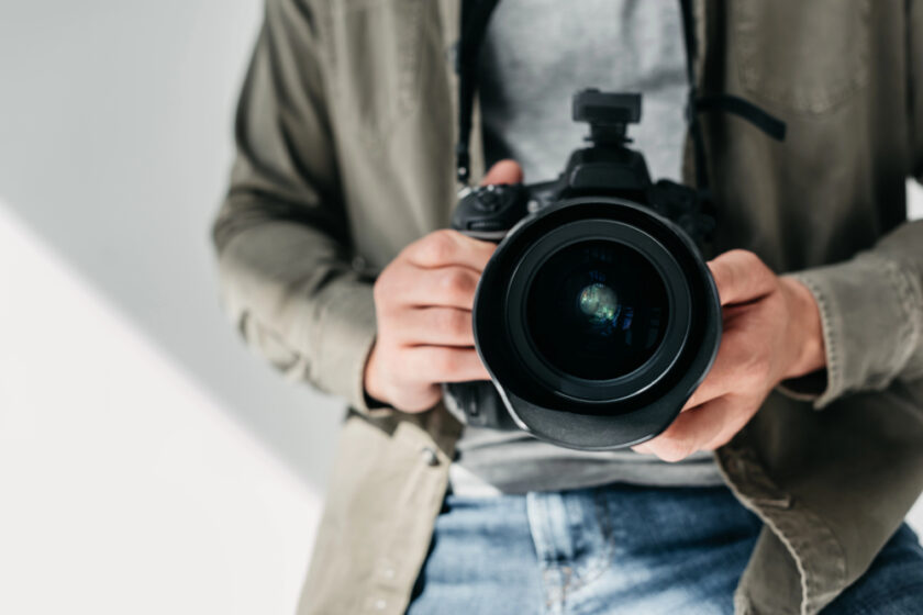 A person wearing a gray shirt and denim jeans holds a professional camera with both hands, pointing the lens directly toward the viewer.