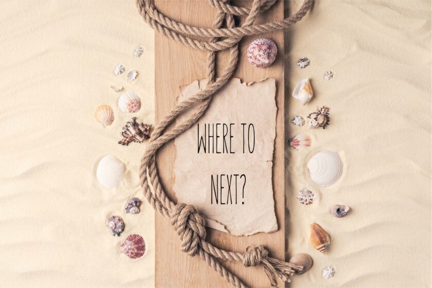 A piece of parchment with the words WHERE TO NEXT? lies on sand, surrounded by seashells and a thick knotted rope atop a wooden plank, evoking a beach or nautical theme.