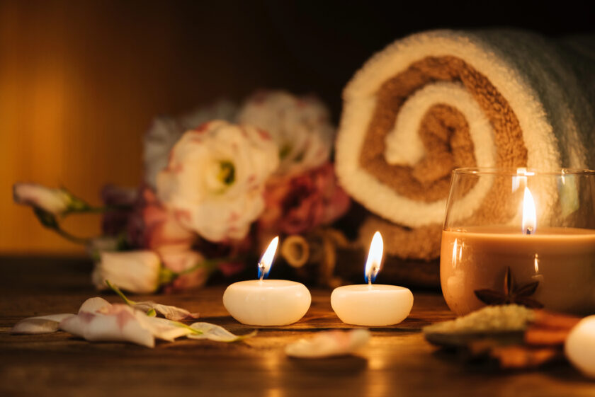A cozy spa setting with two lit tea light candles, a rolled towel, a glass candle, scattered flower petals, and soft-focus flowers in the background on a wooden surface.