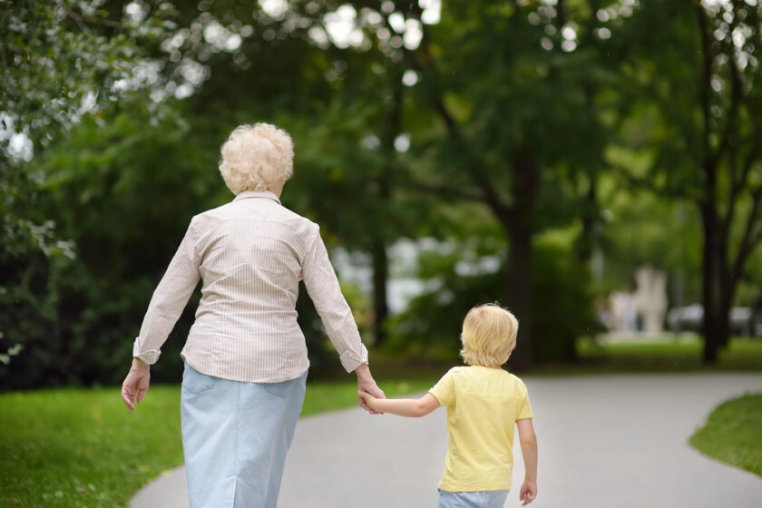 An elderly woman and a young child holding hands walk along a paved path in a green, tree-filled park. The camera captures them from behind as they stroll together.