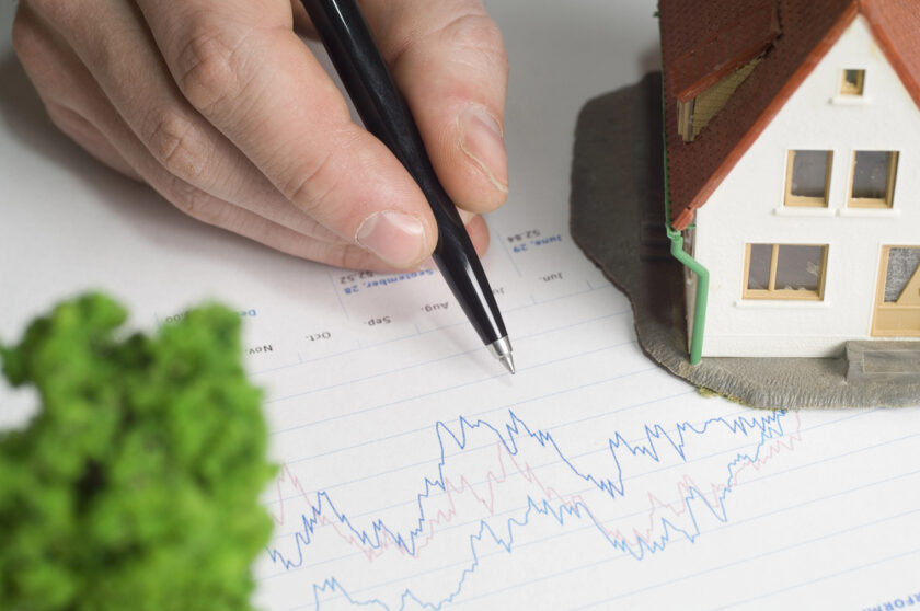A hand holding a pen points to a graph with fluctuating lines on paper, next to a model house and a small green tree, representing real estate market analysis or investment trends.