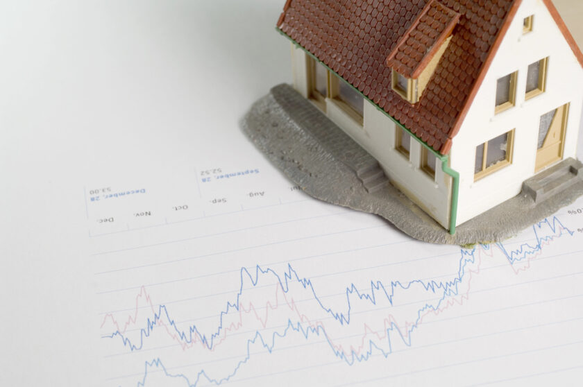A small model house sits on top of financial charts with fluctuating lines, representing data analysis related to real estate or housing market trends.