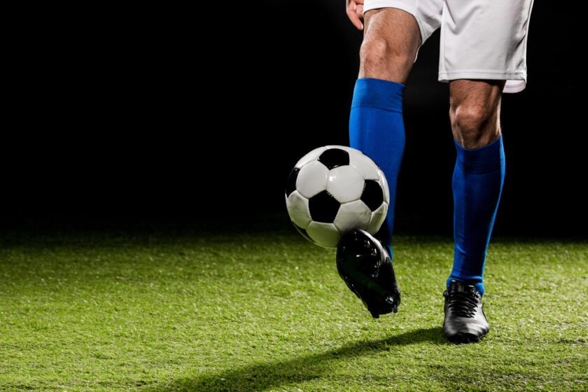 A soccer player wearing blue socks and white shorts is about to kick a black and white soccer ball on a green field, with a dark background.