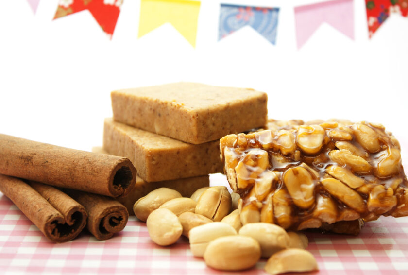 Cinnamon sticks, peanuts, peanut brittle, and three blocks of paçoca are arranged on a pink checkered surface, with colorful paper flags hanging in the background.