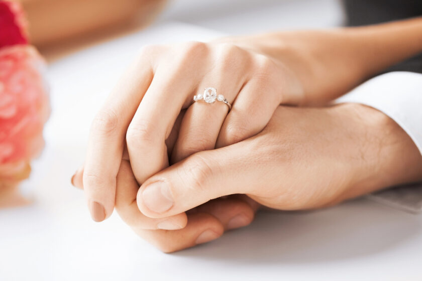 A close-up of two hands gently holding each other, with one hand wearing an engagement ring featuring a diamond. The background is softly blurred.