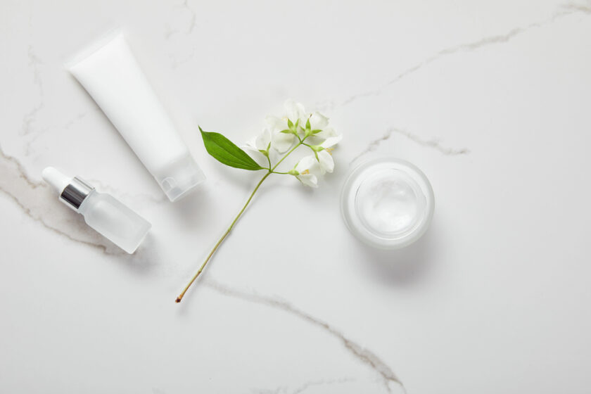 Skincare products and a white flower on a white marble surface; includes a dropper bottle, a tube, and an open jar of cream.