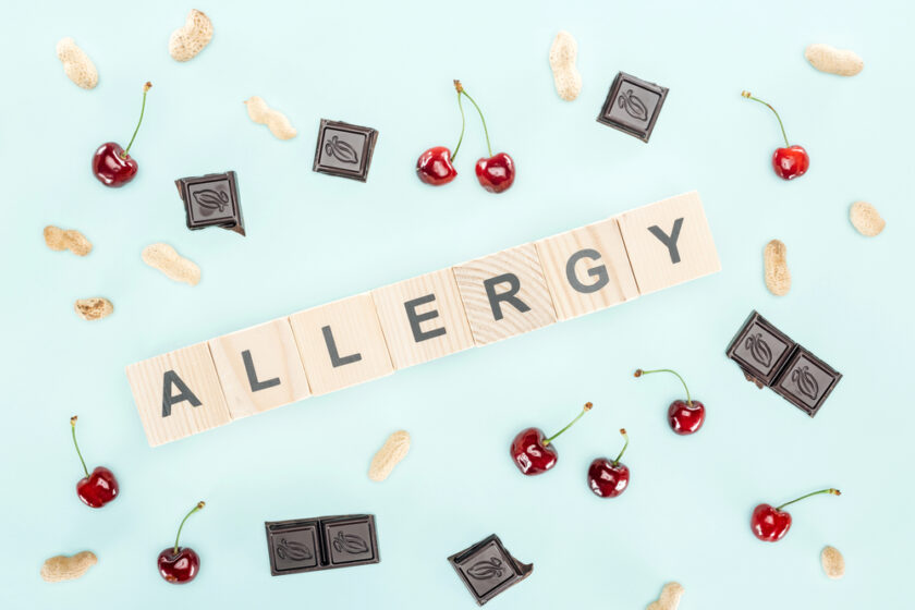 Wooden letter tiles spelling ALLERGY are surrounded by peanuts, cherries, and pieces of dark chocolate, all scattered on a light blue background.