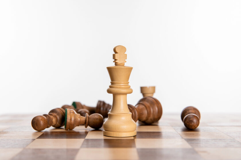 A white queen chess piece stands upright on a chessboard, while several brown chess pieces lie toppled in the background on the board.