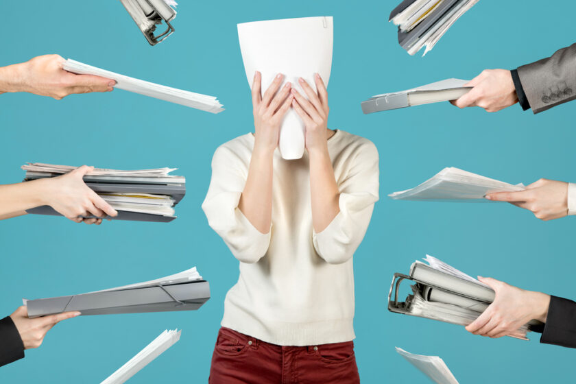 A person covers their face with papers while surrounded by hands holding stacks of documents and folders, suggesting stress or being overwhelmed with work, on a blue background.